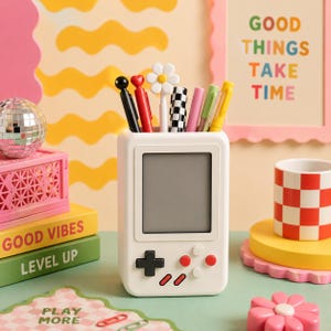 May include: A white desk organiser shaped like a handheld video game console with a blank screen, holding several pens including one with a white daisy topper, a black and white checkered pen, and solid coloured pens in black, red, pink, and yellow.