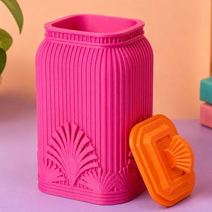 May include: A bright pink rectangular storage canister with vertical ribbed textures and decorative fan-shaped motifs at the base. The canister features an open top and is accompanied by a separate, matching orange lid with a fan-shaped handle.