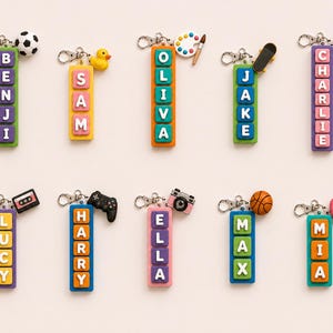 May include: A selection of keyrings with names spelled out in colourful letter blocks. Each keyring has a charm, such as a football, duck, paint palette, skateboard, chocolate, cassette tape, games controller, camera, basketball, and heart-shaped charm.
