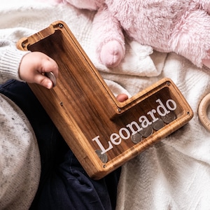 May include: A wooden piggy bank shaped like the letter L with the name "Leonardo" engraved on the side. The piggy bank is filled with coins and a child is putting more coins in.