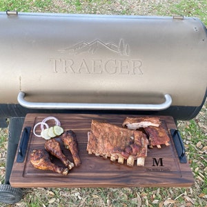 May include: A wooden cutting board with a brown finish, featuring a Traeger grill logo, a personalized inscription "The Miller Family", and a variety of grilled foods, including ribs and chicken legs.
