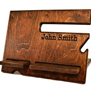 May include: A dark brown wooden phone stand with two slots for phones. The stand has a cutout for charging cables and is personalized with the name "John Smith".