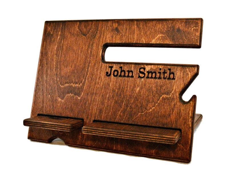 May include: A dark brown wooden phone stand with two slots for phones. The stand has a cutout for charging cables and is personalized with the name "John Smith".