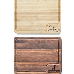 May include: Two wooden cutting boards, one maple and one walnut. The maple board has a rounded corner with the text "The Jacksons Est. 2023". The walnut board has a rounded corner with the letter "J" and the text "The Jackson Family".