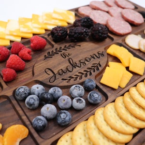 May include: A wooden serving board with multiple compartments for snacks. The board is engraved with "The Jacksons" in a decorative font. The compartments contain cheese, crackers, grapes, berries, and sliced meat.