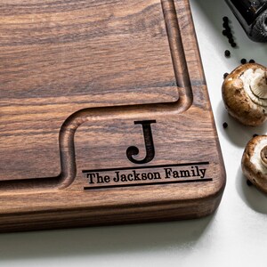 May include: A personalized wooden cutting board with a dark brown finish. The board has a groove around the edge and a smaller groove in the center. The board is engraved with the letter "J" and the text "The Jackson Family".