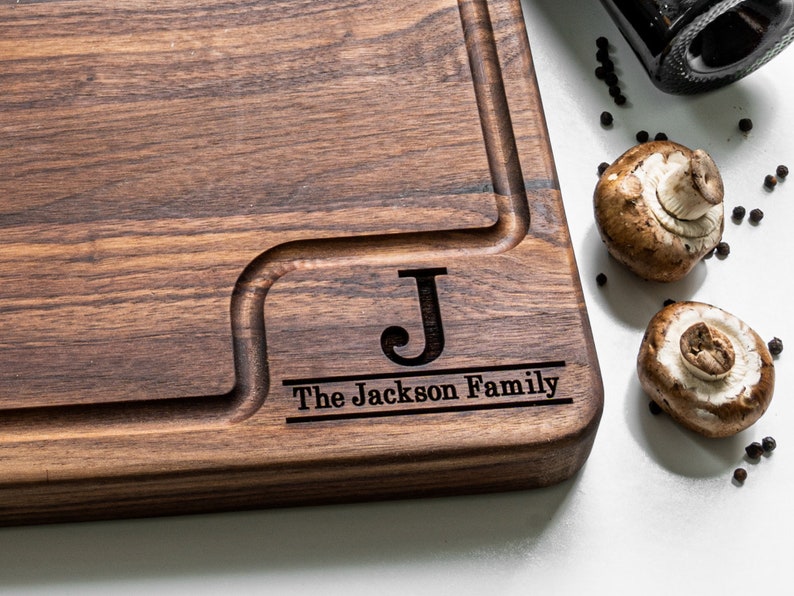 May include: A personalized wooden cutting board with a dark brown finish. The board has a groove around the edge and a smaller groove in the center. The board is engraved with the letter "J" and the text "The Jackson Family".