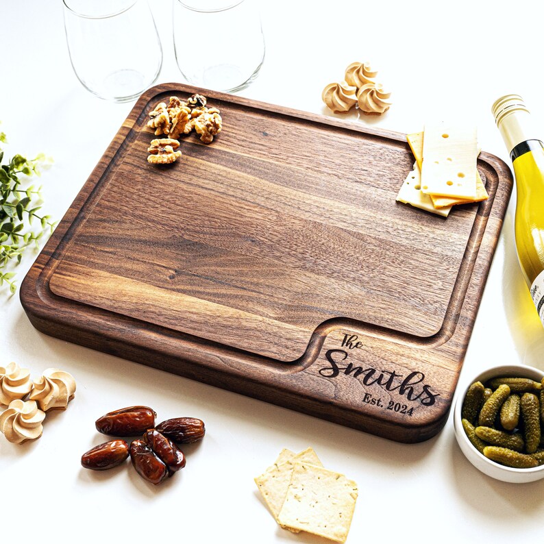 May include: A large, rectangular wooden cutting board with a natural wood grain finish. The board has a raised edge and a carved design in the lower right corner that reads "The Smiths Est. 2024".