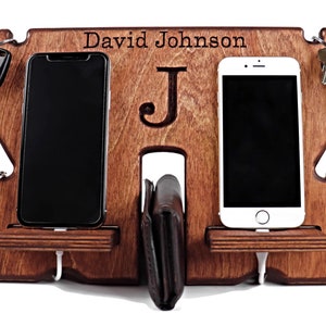 May include: A wooden phone and key holder with a personalized name, "David Johnson", and the initial "J" engraved on it. The holder has two slots for phones, a space for a wallet, and hooks for keys.