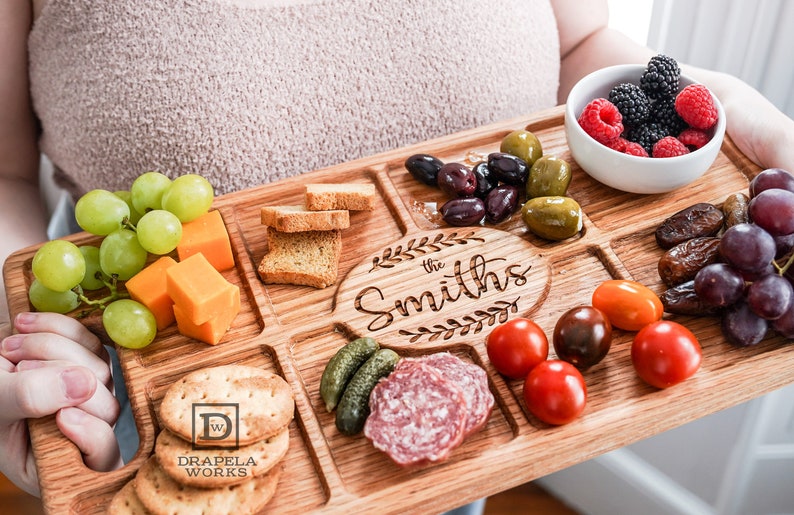 Someone holding a Red Oak Charcuterie Board with Fruit, Ham, Crackers and Cheese on top of the charcuterie