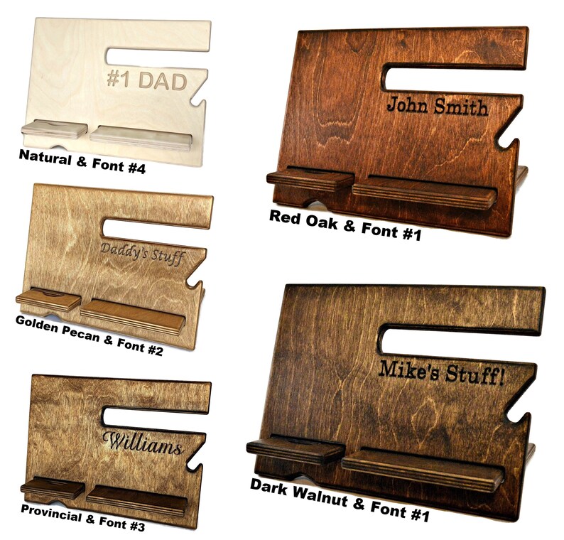 May include: Four wooden phone stands with different wood finishes and engraved text. The stands have two shelves for holding phones and other items. The wood finishes are natural, red oak, golden pecan, and dark walnut. The engraved text includes "#1 DAD", "John Smith", "Daddy's Stuff", "Mike's Stuff!", and "Williams".