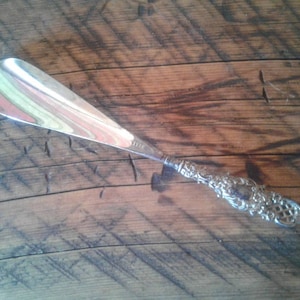 May include: A silver shoe horn with an ornate, detailed handle. The horn has a long, curved shape with a smooth, reflective surface. The handle features intricate floral and scrollwork designs.