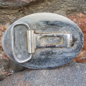 Puede incluir: Hebilla de cintur&oacute;n ovalada de metal desgastado con marco plateado y barra central. La hebilla se encuentra sobre un fondo de rocas rugosas y multicolores, lo que sugiere un tema al aire libre o r&uacute;stico.