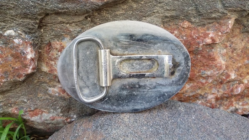 Puede incluir: Hebilla de cintur&oacute;n ovalada de metal desgastado con marco plateado y barra central. La hebilla se encuentra sobre un fondo de rocas rugosas y multicolores, lo que sugiere un tema al aire libre o r&uacute;stico.