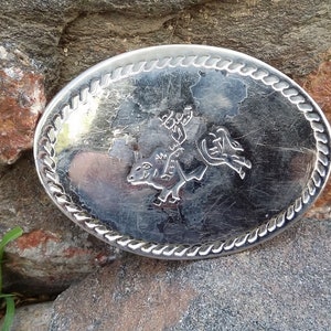 Puede incluir: Hebilla de cintur&oacute;n ovalada plateada con un borde en forma de cuerda. La hebilla presenta un dise&ntilde;o detallado de un jinete de rodeo sobre un bronco. La hebilla est&aacute; sobre un fondo de piedra natural.