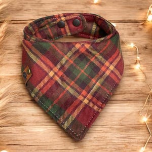 Könnte beinhalten: Ein kariertes Hunde-Bandana in Rot-, Grün- und Gelbtönen. Der Stoff ist mit einem klassischen Karomuster versehen. Das Bandana hat einen Druckknopfverschluss und ein kleines Lederetikett. Der Hintergrund umfasst Weizenähren und Kürbisse.