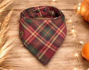 May include: A plaid dog bandana in shades of red, green, and yellow. The fabric is patterned with a classic plaid design. The bandana has a snap closure and a small leather tag. The background includes wheat stalks and pumpkins.