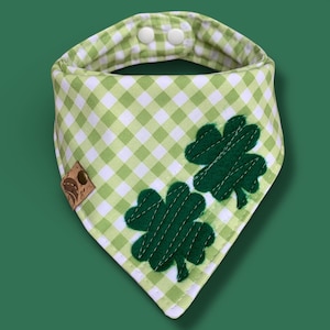 May include: A green and white gingham dog bandana with two dark green felt shamrocks. The bandana has a snap closure and a small brown leather tag. The background is a solid green.