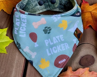 May include: A light blue dog bandana with the text "PLATE LICKER" and illustrations of bones, paw prints, and a football. The bandana has a reversible design with a patterned green fabric on the back and a brown leather accent.