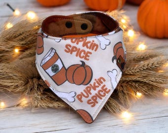 May include: A dog bandana with a cream background featuring a pumpkin spice theme. The bandana has a brown plaid lining and is decorated with pumpkins, coffee cups, and bone illustrations. The text "Pupkin Spice" is printed on the bandana.