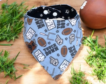 May include: A light blue dog bandana with a football theme. The bandana features brown footballs, play diagrams, and the words "Touch Down." The reverse side is black with white stars. Two white poppers secure the bandana.