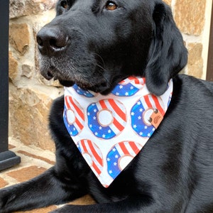 May include: A black Labrador Retriever wearing a white bandana with a red, white, and blue donut pattern. The bandana has a brown leather patch with a small, white, cartoon dog face.