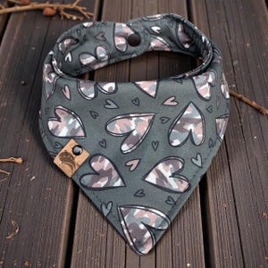 May include: A gray dog bandana with a heart pattern in camouflage colors of white, gray, and pink. The bandana has a snap closure and a small leather tag with a logo. The background is a wooden surface.