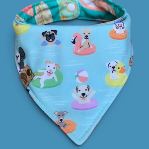 May include: A light blue dog bandana with a pattern of dogs on floats in a pool. The bandana has a snap closure and a reversible design with a colorful striped pattern on the reverse side.