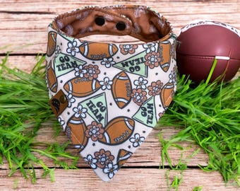 May include: A beige dog bandana with a football and flower pattern, and the words "GO TEAM" in green triangles. A brown football sits next to the bandana on a wooden surface with green grass.