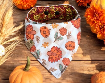 May include: A white bandana with a Thanksgiving theme. It features a checkered pattern with images of turkeys, pumpkins, and fall flowers. The bandana has a brown fabric trim and snaps on the back.