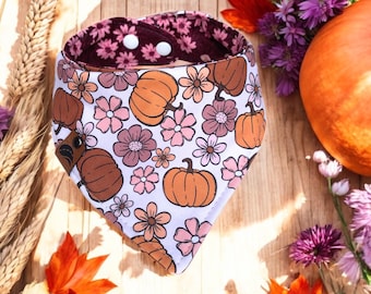 May include: A white bandana with a fall-themed pattern of pumpkins and flowers in shades of brown, orange, and pink. The bandana has a burgundy floral lining and white snap closures. The bandana is on a wooden surface.