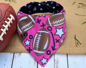 May include: A pink dog bandana with a football theme. The bandana features brown American footballs, white stars, and black play diagrams on a pink background. The reverse side is black with white stars. A brown football is visible on the left.