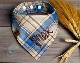 May include: A plaid dog bandana in blue, tan, and brown with the name "MAX" embroidered in brown thread. The bandana has a snap closure and a small leather tag. Wheat stalks are in the background.