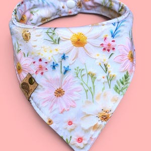 May include: A floral dog bandana featuring a white background with pink, yellow, and blue flower patterns. The bandana has a snap closure and a small brown leather tag.