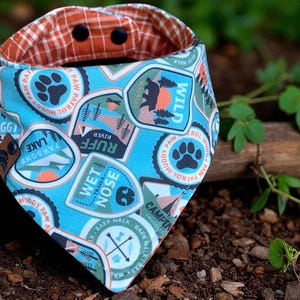 Puede incluir: Un pañuelo azul para perros con varios parches con temas de actividades al aire libre, que incluyen huellas de patas, un oso y las palabras "Wild" y "Ruff River". El pañuelo tiene un diseño reversible con un patrón a cuadros naranja y marrón. Se asegura con dos broches negros.