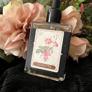 May include: A clear glass bottle with a black dropper top and a label that says "COSETTE". The label features a vintage illustration of a rose with a key and a ribbon.
