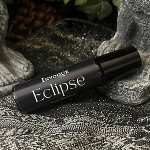 May include: A black roll-on perfume bottle with the word "Eclipse" in white script. The bottle is labelled "Invouque" and "Higher Perfumer". The background is a textured, dark fabric with a stone and foliage.