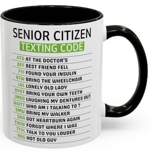 Op de afbeelding: Een witte keramische mok met een zwarte binnenkant en handvat. De mok heeft de tekst "SENIOR CITIZEN TEXTING CODE" in groen en zwart, samen met een lijst met sms-afkortingen. Een humoristisch cadeau-idee.
