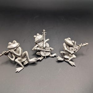 May include: Three silver-colored frog figurines in a row, each playing a different instrument. One frog plays a flute, another a cello, and the third a violin. The frogs are detailed with realistic features and are set against a dark background.