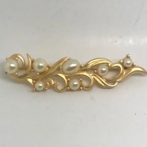 Puede incluir: Broche dorado con un diseño fluido y orgánico. La broche está adornada con varias perlas de imitación de diferentes tamaños, lo que añade un toque clásico. La pieza tiene una apariencia delicada y elegante, adecuada para ocasiones formales.