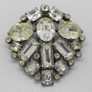 May include: A vintage brooch with a silver-tone metal setting. The brooch features a central oval-cut clear stone, surrounded by various rectangular, square, and round clear and light yellow stones. A classic piece of jewelry.