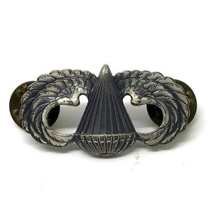 May include: A silver-toned military badge featuring a central conical design with a textured surface, flanked by stylized wings. The badge has a pin backing and is set against a white background.