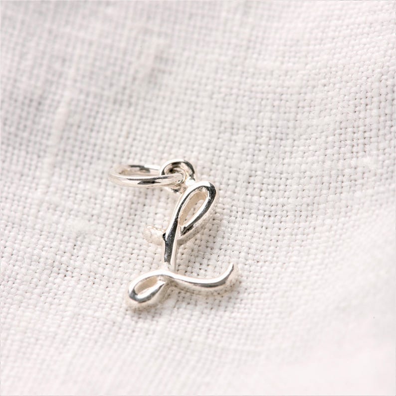 Może przedstawiać: Srebrny charms w kształcie litery "L" w kursywie. Charms ma małą pętelkę u g&oacute;ry do przymocowania do naszyjnika lub bransoletki. Charms jest prezentowany na białej, teksturowanej powierzchni.