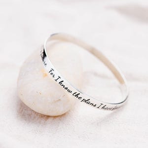 May include: A silver bangle bracelet with the inscription "For I know the plans I have for you" in black script. The bracelet is resting on a smooth, light-colored stone, showcasing its elegant design.