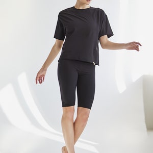 K&ouml;nnte beinhalten: Eine Frau tr&auml;gt ein schwarzes T-Shirt mit kurzen &Auml;rmeln und schwarze Radlerhosen. Sie steht auf einem wei&szlig;en Hintergrund.