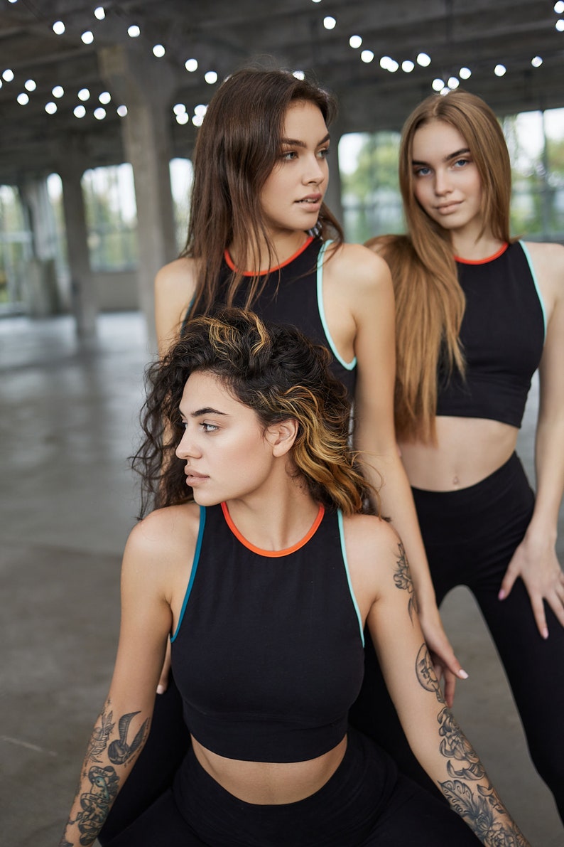 May include: Three women wearing black athletic tops with colorful trim. The woman in the front has a tattoo on her arm.