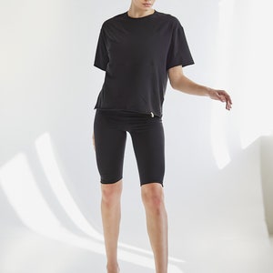K&ouml;nnte beinhalten: Eine Frau tr&auml;gt ein schwarzes T-Shirt mit kurzen &Auml;rmeln und schwarze Radlerhosen. Sie steht auf einem wei&szlig;en Hintergrund.