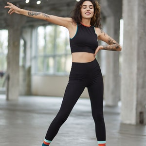 May include: A woman wearing a black crop top and black leggings with colorful stripes at the bottom. She is wearing black sneakers and has her arms outstretched.