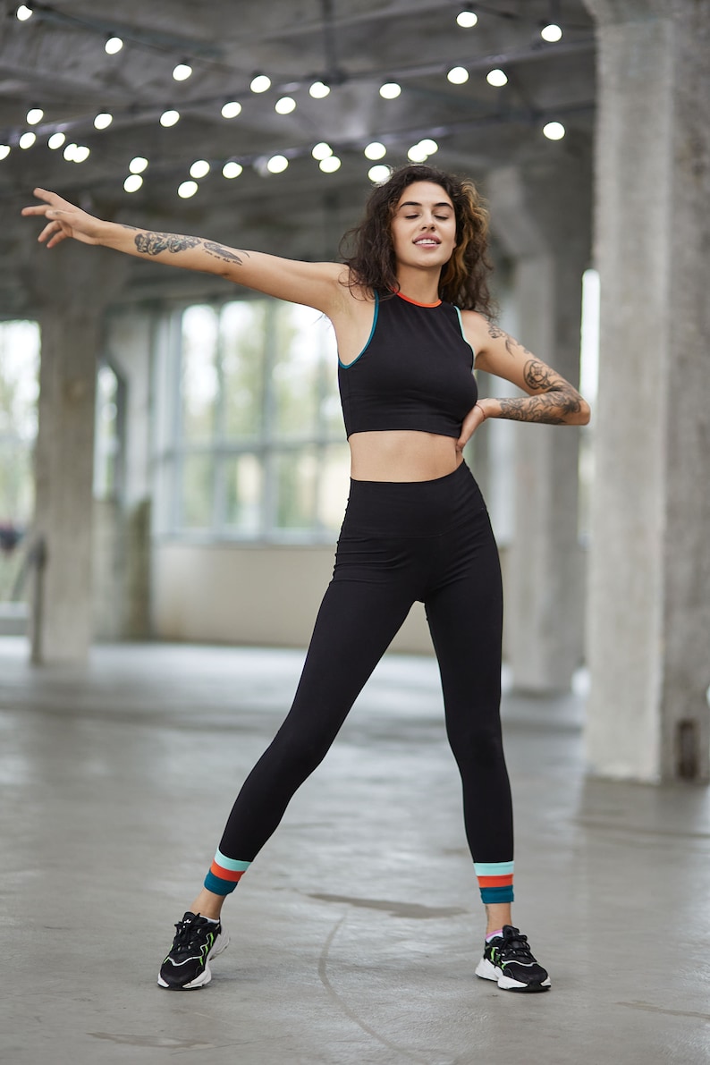 May include: A woman wearing a black crop top and black leggings with colorful stripes at the bottom. She is wearing black sneakers and has her arms outstretched.