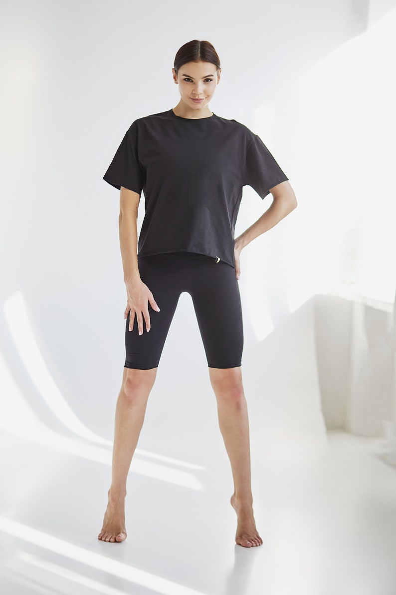 K&ouml;nnte beinhalten: Eine Frau tr&auml;gt ein schwarzes T-Shirt mit kurzen &Auml;rmeln und schwarze Radlerhosen. Sie steht auf einem wei&szlig;en Hintergrund.